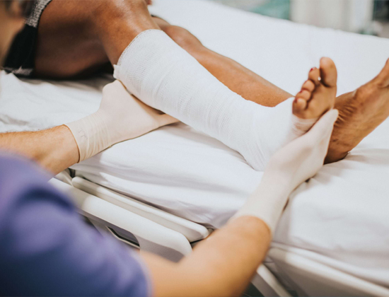 A healthcare professional wearing gloves carefully handling a patient’s leg with a bandaged wound in a clinical setting.