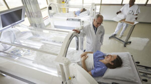 A medical facility with a patient lying inside a hyperbaric oxygen therapy chamber, a doctor interacting with another patient, and a staff member reviewing documents in the background.